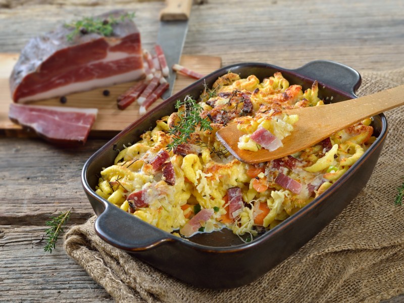 schwarte Auflaufform mit Spätzle-Auflauf auf einem dunklen Tisch und Schinken im Hintergrund