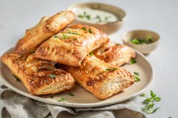 mehrere Spinat-Blätterteig-Taschen mit Feta auf einem Teller, im Hintergrund eine Schale mit Dip und eine mit Kräutern.