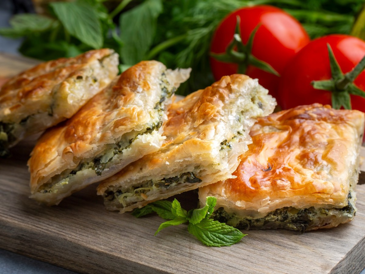 4 Stücke Spinat-Börek mit Feta auf einem Brett, dahinter Tomaten und frischer Spinat.