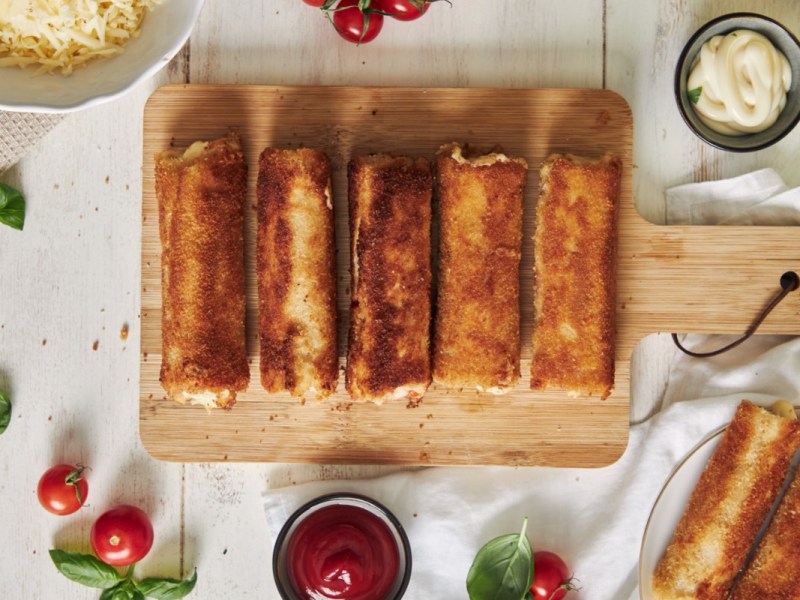 Fünf Toast-Röllchen auf einem Holzbrett. Drumherum stehen Schalen mit Dip und frische Zutaten.