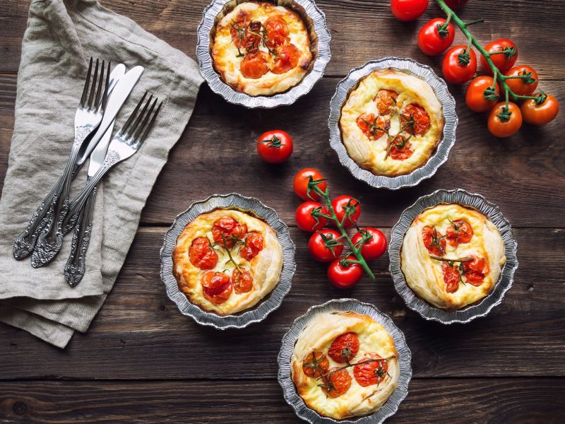 Tomaten-Ziegenkäse-Tartes in kleinen Formen stehen auf einem Brett. Frische Tomaten und Besteck liegen als Deko daneben.