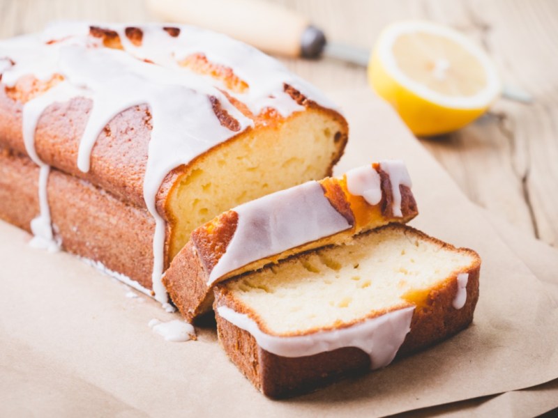 Ein Zitronen-Mascarpone-Kuchen, der an einer Seite schon in Stücke geschnitten wurde. Im Hintergrund liegt eine halbe Zitrone.