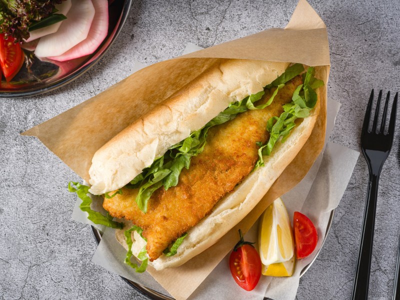 Backfischbrötchen in einem Pergamentbeutel. Daneben Messer und Gabel, Tomate und Zitrone.