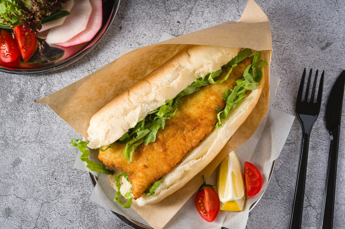 Backfischbrötchen in einem Pergamentbeutel. Daneben Messer und Gabel, Tomate und Zitrone.