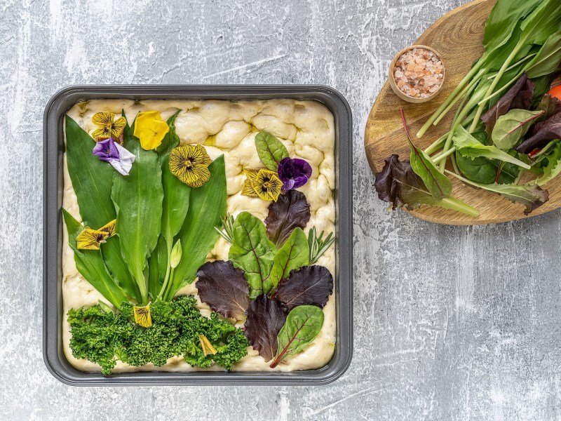 Bärlauch-Focaccia, auf die eine Wiese mit Gemüse wie Bärlauch und Tomaten gelegt wurde, ungebacken, daneben ein Brett mit frischem Gemüse, Draufsicht.