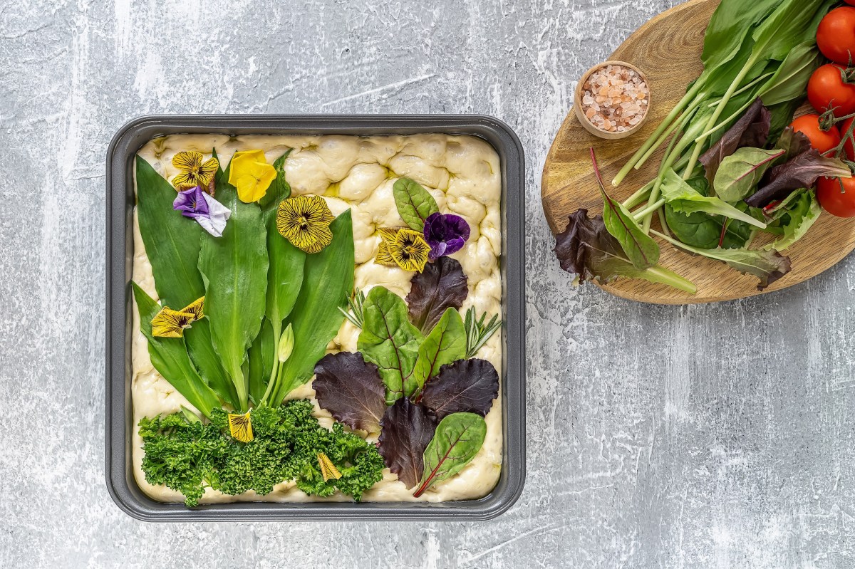 Bärlauch-Focaccia, auf die eine Wiese mit Gemüse wie Bärlauch und Tomaten gelegt wurde, ungebacken, daneben ein Brett mit frischem Gemüse, Draufsicht.