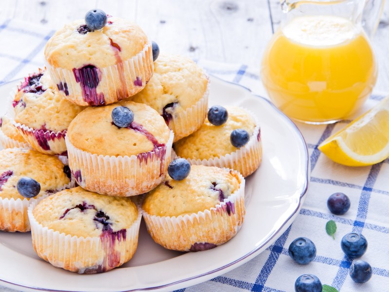 Ein Teller mit mehreren Blaubeer-Buttermilch-Muffins auf kariertem Tischtuch. Kanne mit Zitronensaft daneben.