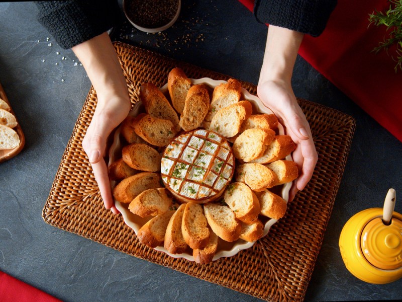 Camembert-Blume in einer Auflaufform, umgeben von Kräuterbaguette. Gehalten wird er von zwei Händen einer Frau