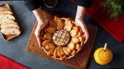 Camembert-Blume in einer Auflaufform, umgeben von Kräuterbaguette. Gehalten wird er von zwei Händen einer Frau