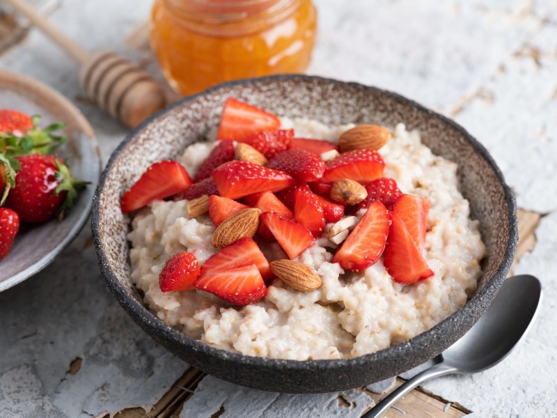 Eine dunkle Schüssel Erdbeer-Porridge, garniert mit Erdbeeren und ganzen Mandeln, daneben ein Teller mit Erdbeeren und ein Löffel.