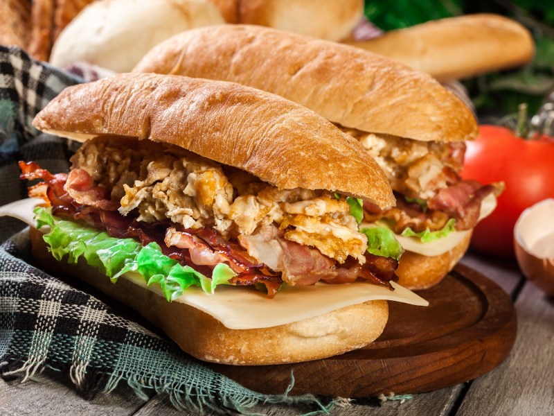 Zwei Frühstücks-Panini auf einem Holzbrett, das auf einer karierten Tischdecke steht. Im Hintergrund sind unscharf eine Tomate und eine Eierschale zu erkennen.