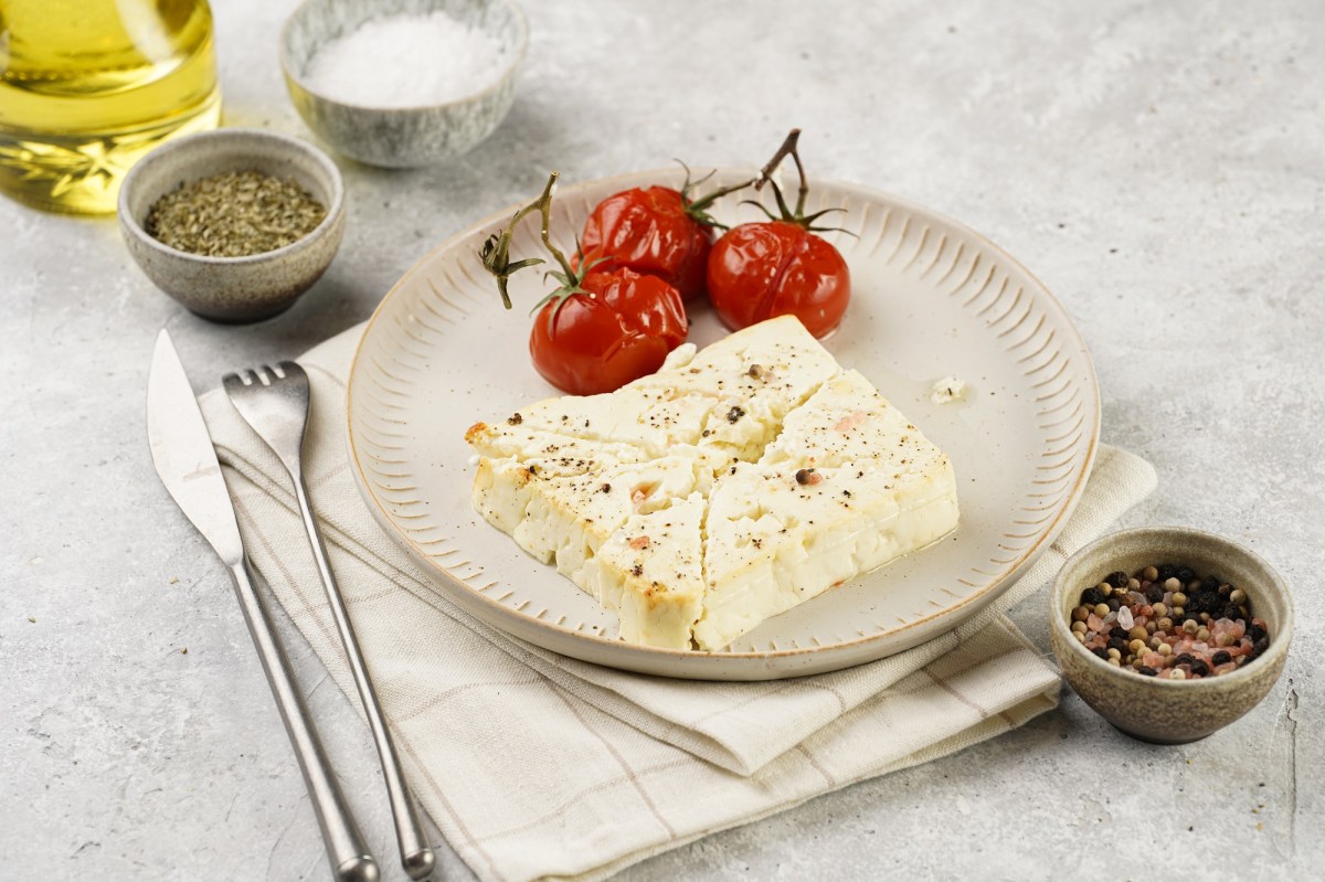 Gebackener Schafskäse auf einem Teller, daneben gebackene Tomaten mit Strunk, Besteck, eine Schale Salz und Pfeffer sowie eine Flasche Öl.