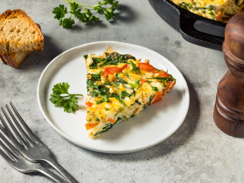 Ein Stück Gemüse-Tortilla auf einem weißen Teller, garniert mit Petersilie. Daneben liegen einige Scheiben Brot und es steht eine Pfeffermühle. Im Hintergrund ist der Rest Tortilla zu erahnen.