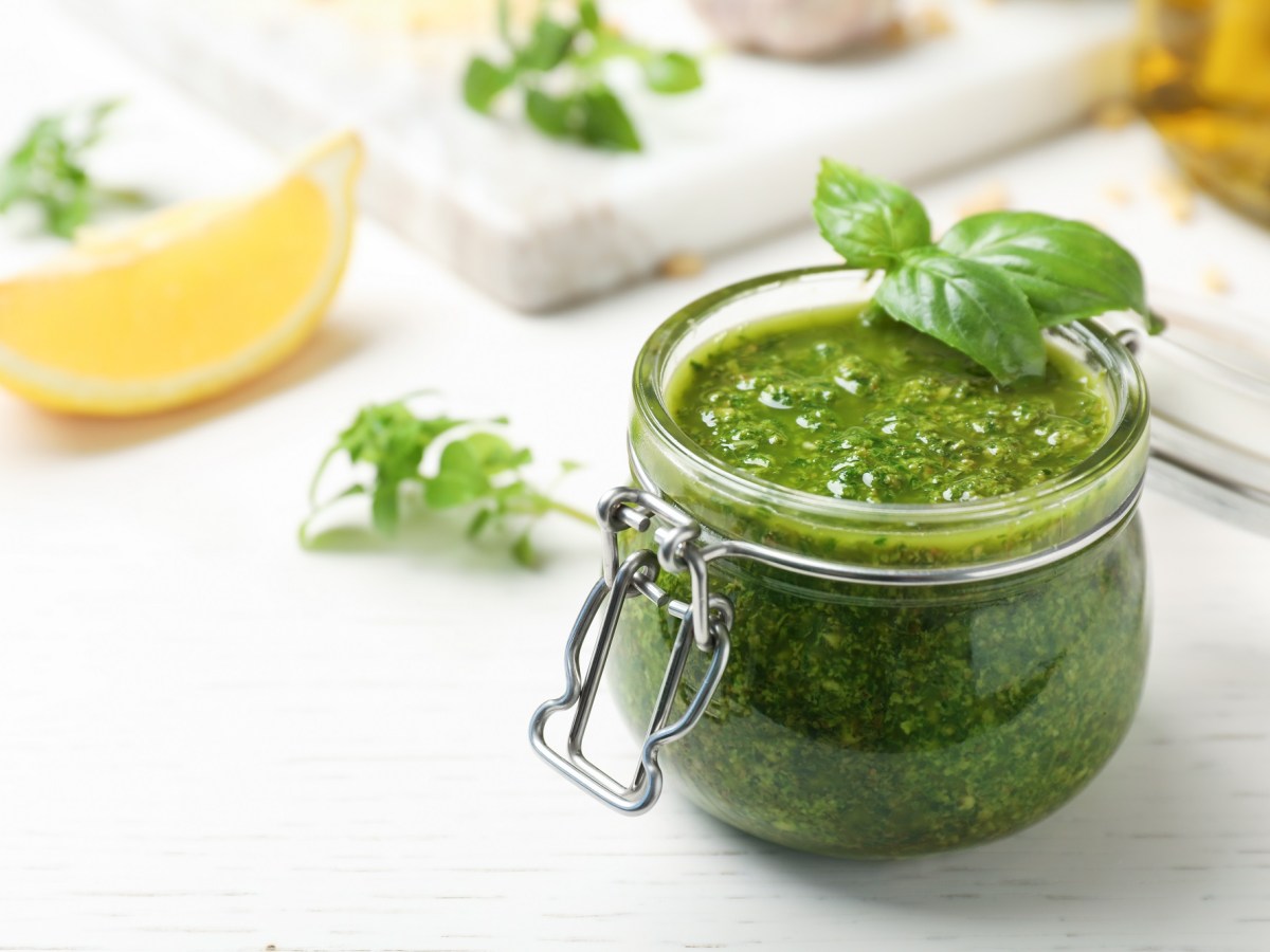 Ein Glas Basilikumpesto, ein grünes Pesto-Rezept, daneben eine Zitronenspalte und Basilikum.