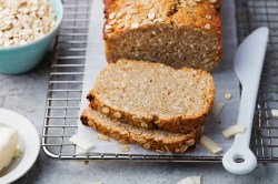 angeschnittenes Haferflocken-Quark-Brot auf einem Brett, daneben eine Schüssel Haferflocken und ein Messer.