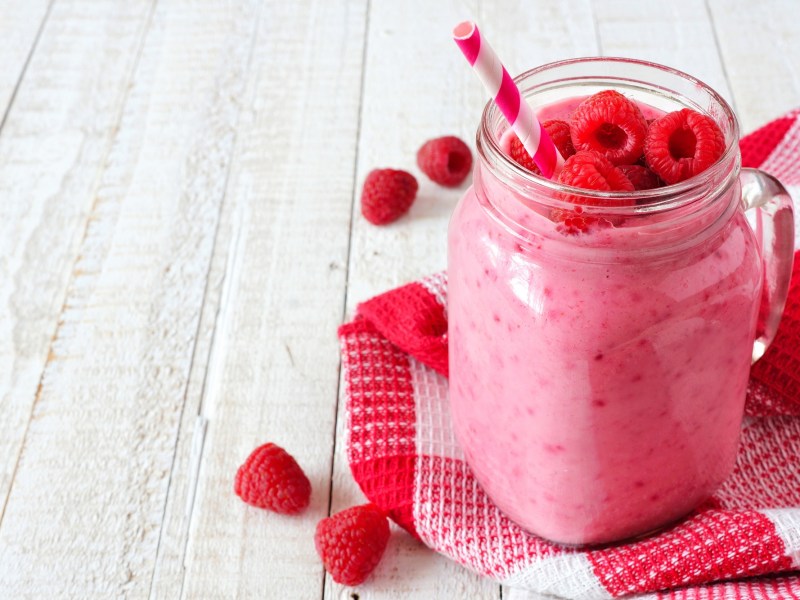 Ein Maison Jar Glas mit Himbeer-Joghurtdrink, garniert mit Himbeeren, darin ein Strohhalm, drumherum frische Beeren und ein kariertes Geschirrtuch.