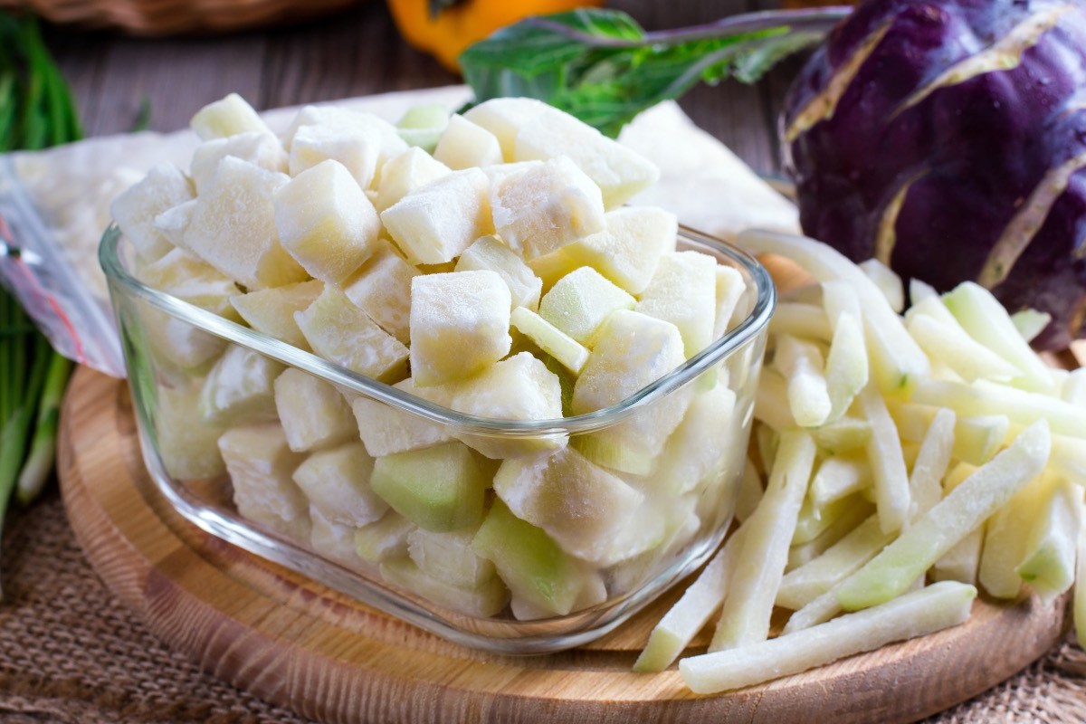 Kohlrabi einfrieren: gefrorene Kohlrabistücke in einer Glasschüssel. Im Hintergrund liegt frisches Gemüse.