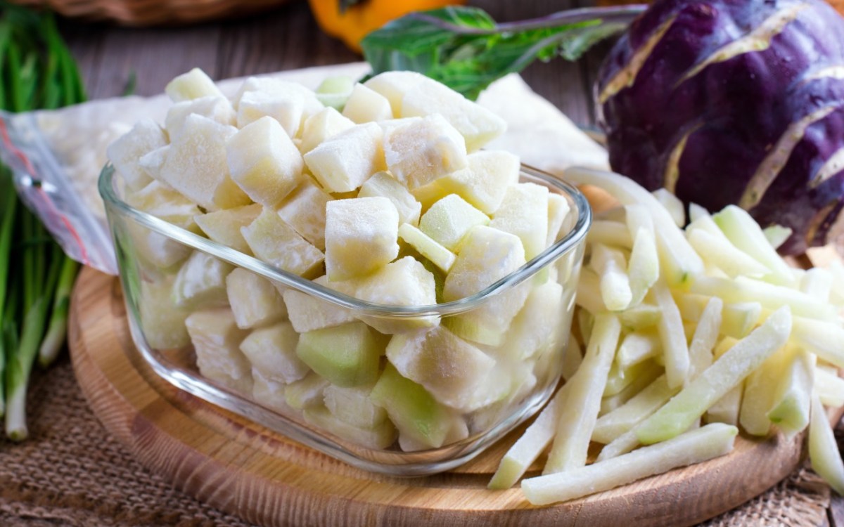 Kohlrabi einfrieren: gefrorene Kohlrabistücke in einer Glasschüssel. Im Hintergrund liegt frisches Gemüse.