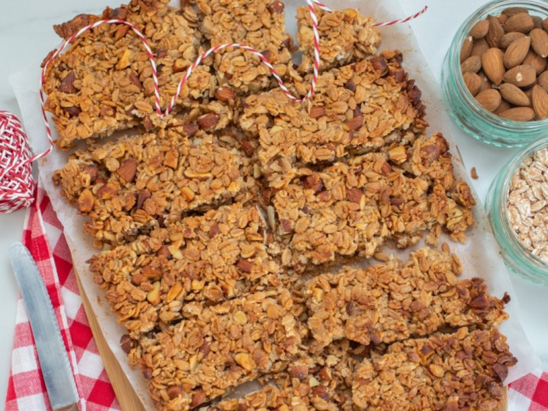 Müslikuchen in mehrere Stücke zerteilt, in der Draufsicht. Daneben stehen Schalen mit Mandeln und Haferflocken.