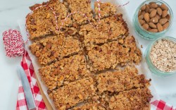 Müslikuchen in mehrere Stücke zerteilt, in der Draufsicht. Daneben stehen Schalen mit Mandeln und Haferflocken.