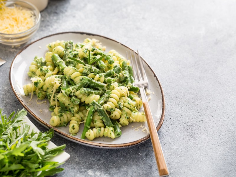 Ein ovaler Teller Nudeln mit Spargel und Rucola, darauf eine Gabel, daneben frischer Rucola und eine Schale Käse.