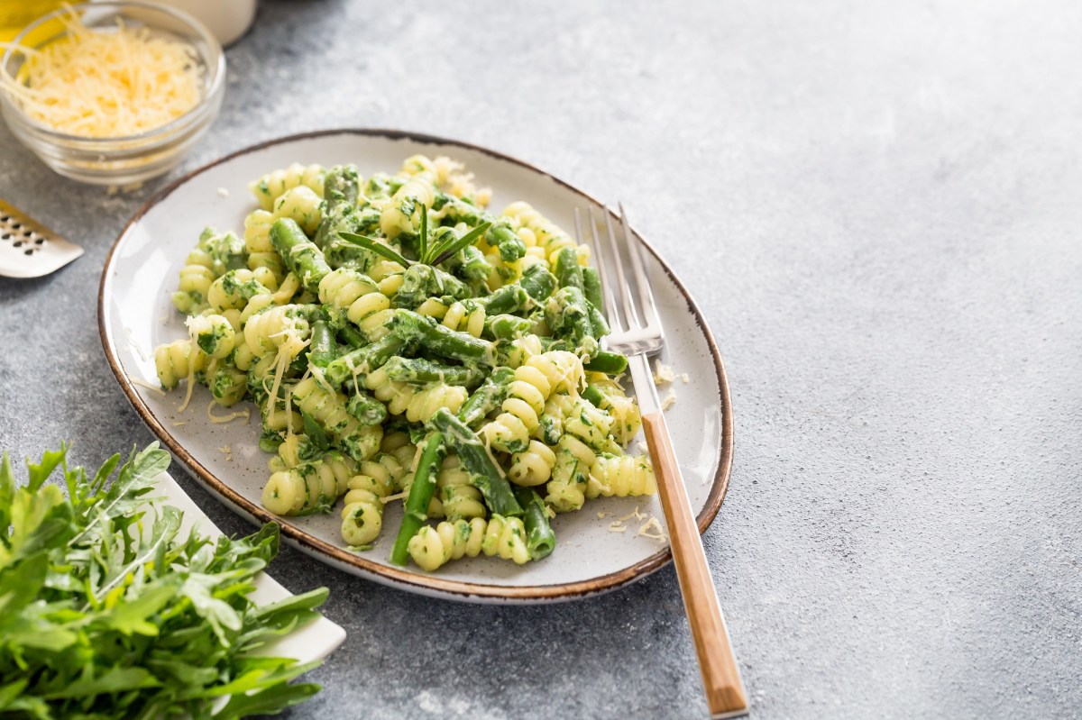 Ein ovaler Teller Nudeln mit Spargel und Rucola, darauf eine Gabel, daneben frischer Rucola und eine Schale Käse.