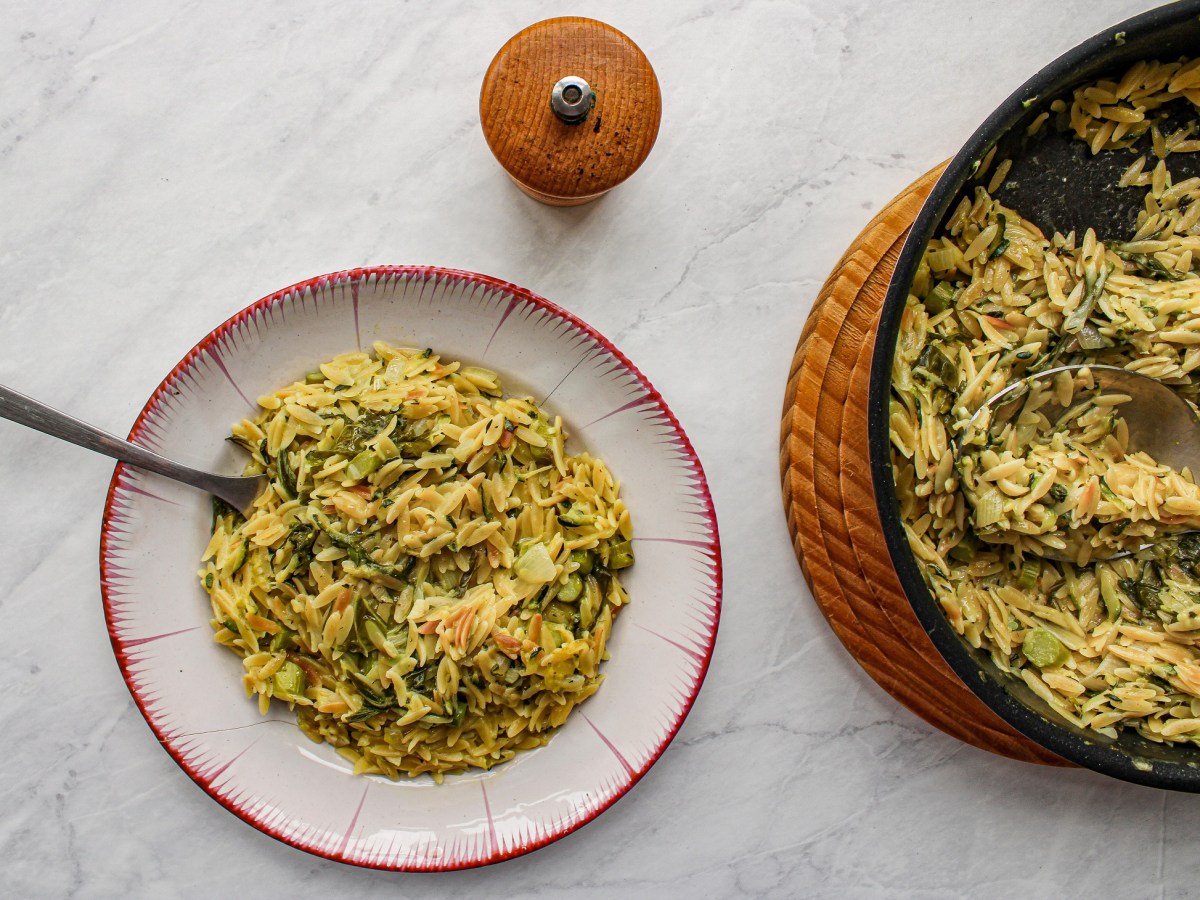 Ein Teller Orzo-Nudeln mit grünem Spargel in der Draufsicht, darin eine Gabel, daneben ein Gewürzstreuer und die Pfanne mit dem Gericht.