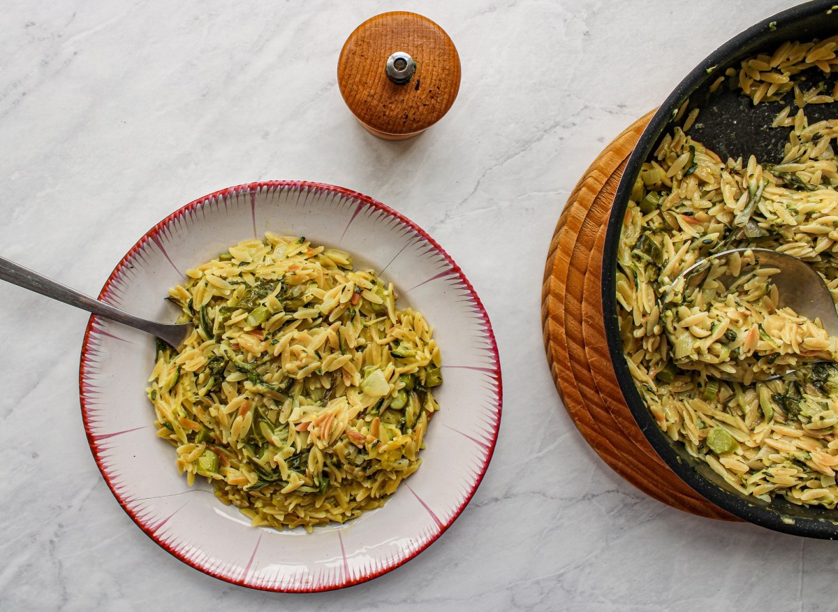 Ein Teller Orzo-Nudeln mit grünem Spargel in der Draufsicht, darin eine Gabel, daneben ein Gewürzstreuer und die Pfanne mit dem Gericht.