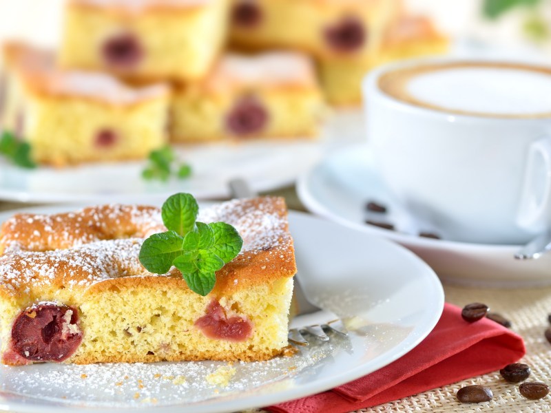 Quark-Kirsch-Kuchen auf einem weißen Teller. Im Hintergrund steht mehr Kuchen, daneben eine Tasse Cappuccino.