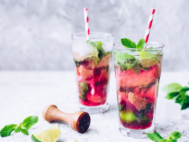 Zwei Gläser Rhabarber-Mojito mit Minze, Limette und Barstößel daneben.