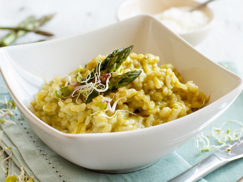 Ein tiefer Teller Risotto mit Sauce hollandaise, grünen Spargelspitzen und Sprossen.