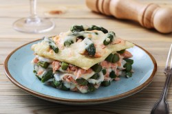 Ein Stück Spargel-Lachs-Lasagne auf einem hellblauen Teller. Daneben liegt eine Gabel und im Hintergrund liegt eine Pfeffermühle.