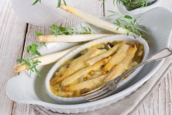 Spargel-Lauch-Auflauf in einer weißen Auflaufform. Daneben Stangen weißer Spargel und Zweige Lavendel.