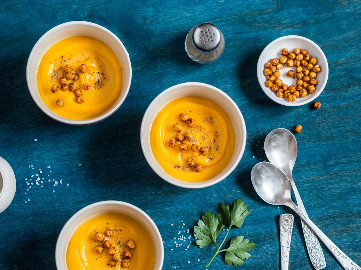 Drei Schalen mit orangefarbener Süßkartoffelsuppe mit gerösteten Kichererbsen und Löffel daneben auf blauem Untergrund.