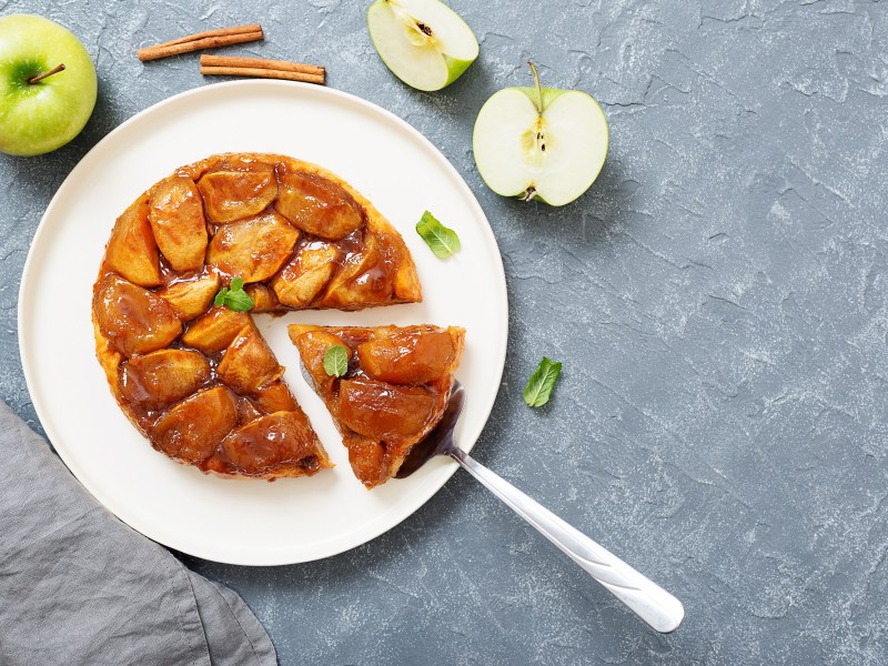 Tarte Tatin auf einem weißen Teller. Es wurde bereits ein Stück abgeschnitten. Daneben liegen angeschnittene grüne Äpfel und ein paar Zimtstangen.