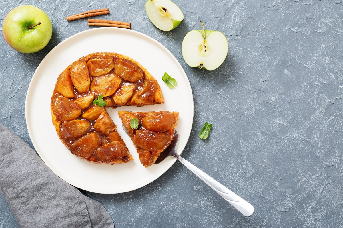 Tarte Tatin auf einem weißen Teller. Es wurde bereits ein Stück abgeschnitten. Daneben liegen angeschnittene grüne Äpfel und ein paar Zimtstangen.