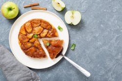 Tarte Tatin auf einem weißen Teller. Es wurde bereits ein Stück abgeschnitten. Daneben liegen angeschnittene grüne Äpfel und ein paar Zimtstangen.