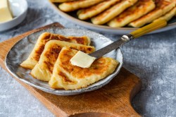 Tattie Scones, serviert mit Butter, auf einem grauen Teller. Im Hintergrund stehen weitere Tattie Scones.