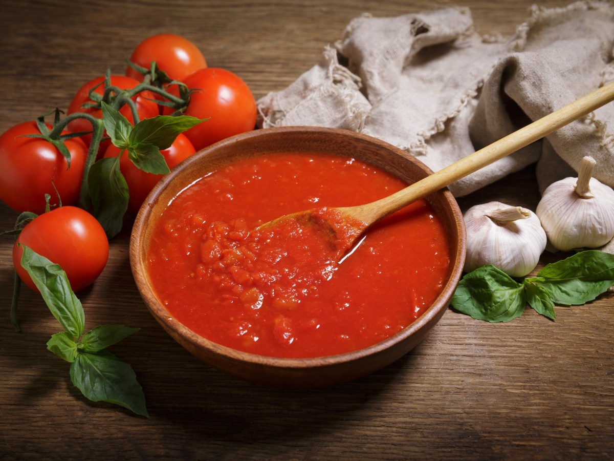 Tomatensoße in einer Schüssel. Daneben Tomaten, Knoblauch und Basilikum.