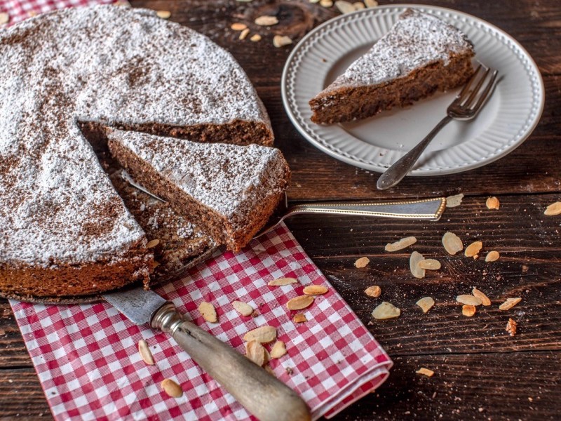 Torta Caprese, italienischer Schoko-Mandel-Kuchen, auf einem weißen Teller. Daneben ein Teller mit einem Stück Kuchen.