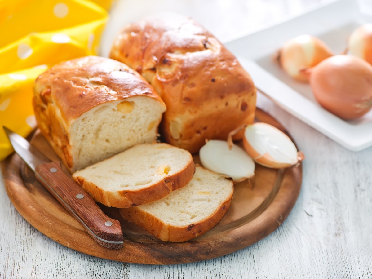 Zwiebelbrot: Zwei Laibe auf einem Holzbrett, eines davon ist angeschnitten. Im Hintergrund liegen Zwiebeln