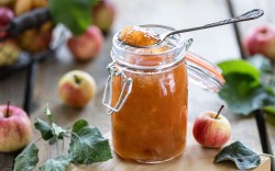 Ein Glas Apfelmarmelade mit Senf, in der ein Löffel steckt. Drumherum liegen Äpfel.