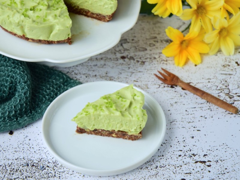 Ein Stück Avocado-Limetten-Käsekuchen auf einem Teller. Im Hintergrund der restliche Kuchen und ein paar gelbe Narzissen.