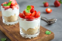 2 Cheesecake im Glas mit Keksboden und Erdbeeren garniert auf einem Brett, im Hintergrund Löffel und Erdbeeren.