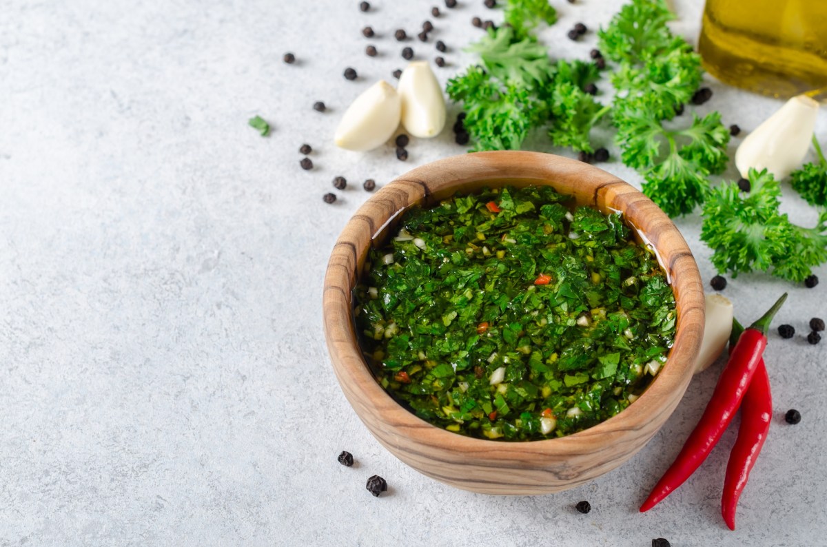 Eine Holzschale Chimichurri, daneben Knoblauchzehen, Chilischoten, Kräuter und Pfefferkörner.