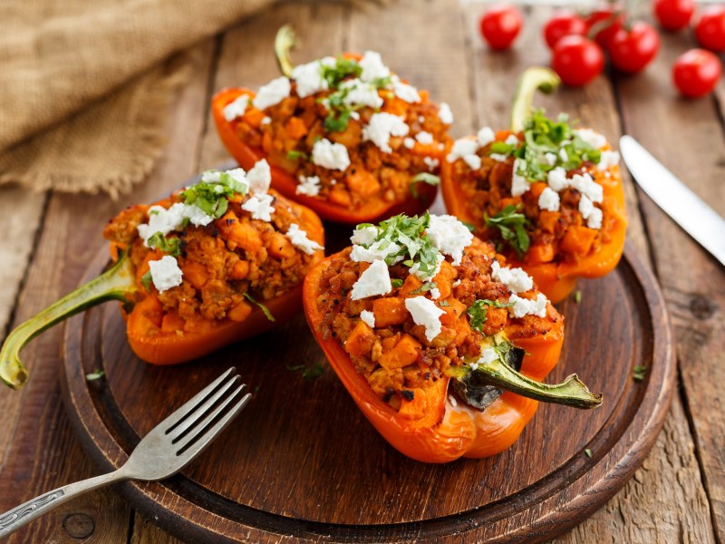 Vier Hälften gefüllte Paprika vom Grill liegen auf einem Holzbrett. Daneben Besteck.