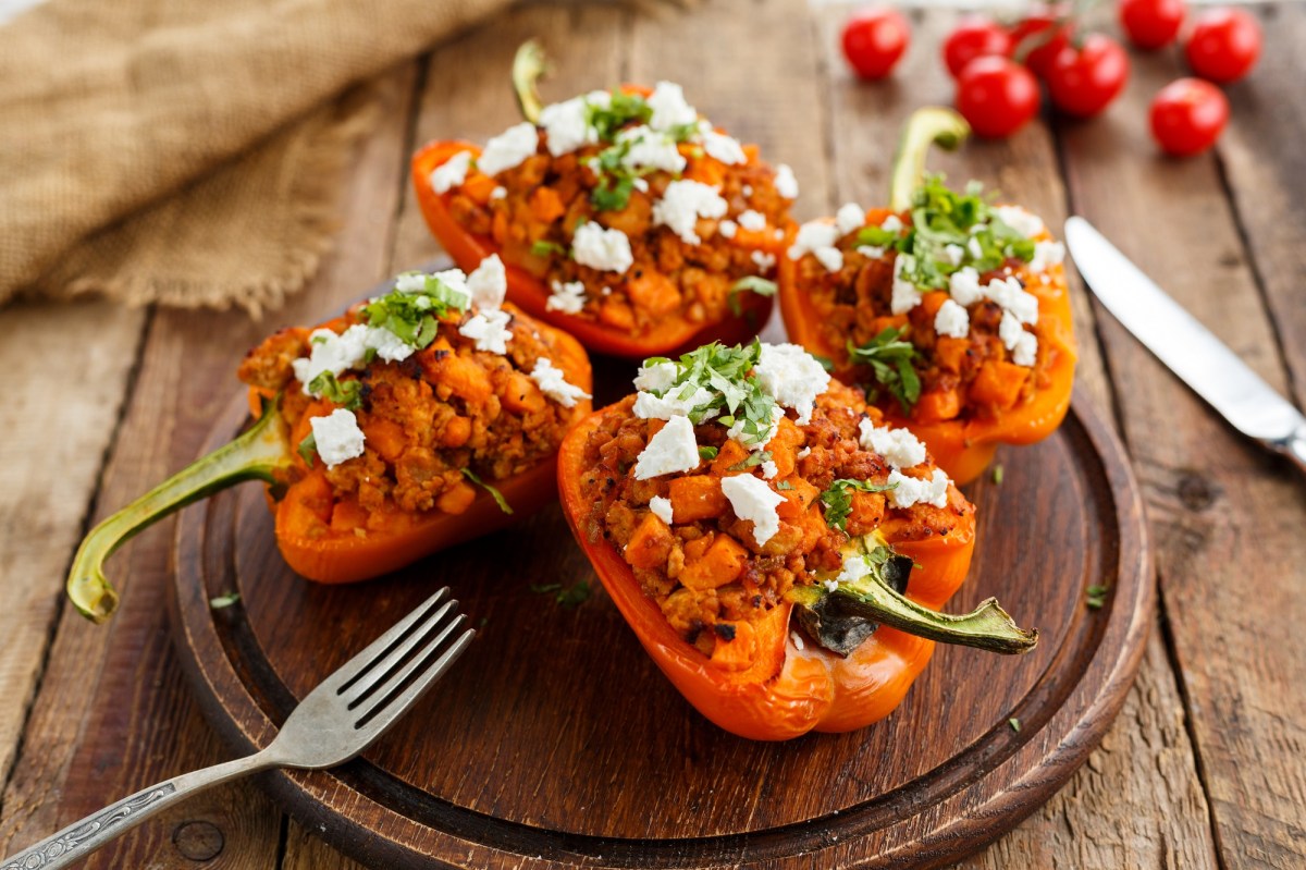 Vier Hälften gefüllte Paprika vom Grill liegen auf einem Holzbrett. Daneben Besteck.