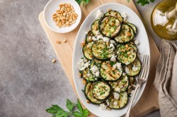 Gegrillte Zucchini in Scheiben mit Feta und Pinienkernen bestreut auf einem ovalen Teller, Draufsicht. Daneben Chiliflocken in einer Schüssel, eine Ölflasche und andere Zutaten.