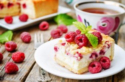 Ein Stück Himbeer-Joghurt-Kuchen auf einem weißen Teller. Dahinter eine Tasse Tee und unscharf der Rest des Kuchens.