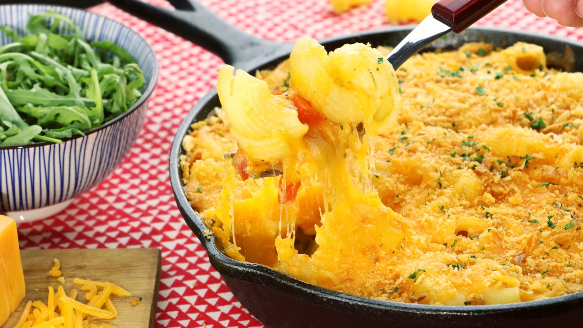 Käsenudeln, serviert in einer gusseisernen Pfanne. Im Hintergrund stehen Tomaten und Semmelbröseln als Deko. Nahaufnahme